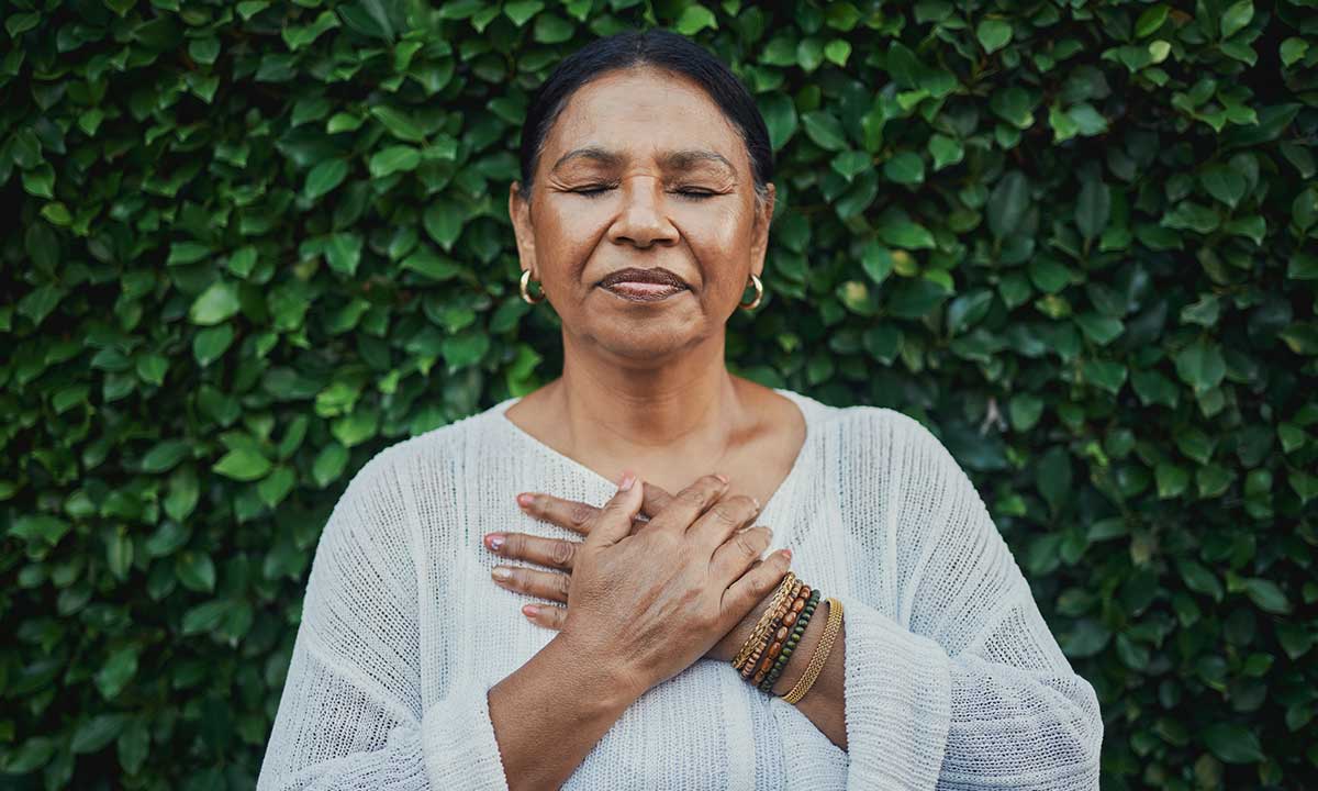 A woman with eyes closed holds her hands over her heart in front of green leaves, expressing a peaceful moment of gratitude.