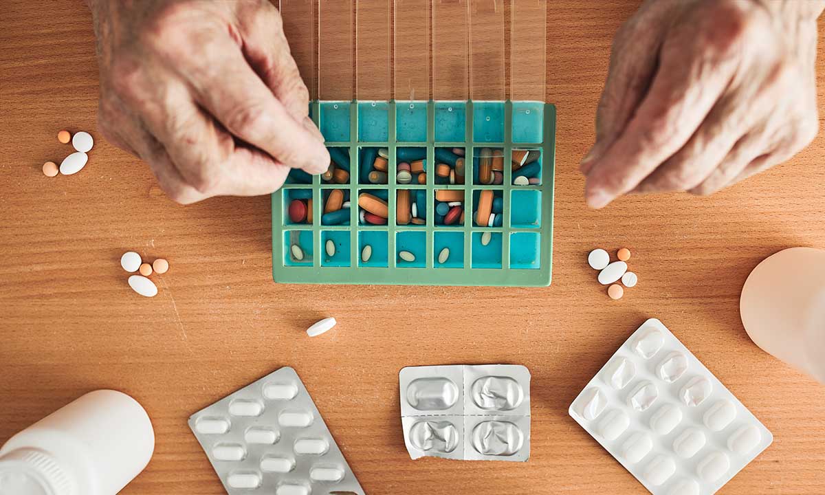 Close-up of an older adult’s hands sorting prescription pills into a weekly medication organizer.