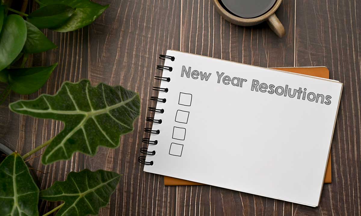 Notebook labeled “New Year Resolutions” with a checklist on a wooden table, surrounded by green plants and a coffee mug.
